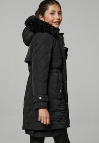 Črna prešita parkas jakna z umetnim krznom na kapuci, zatiči, elastičnim pasom in stranskimi žepi. Ponuja gladko teksturo in prirejen kroj.