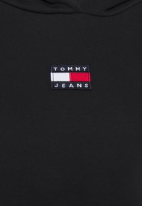Černá mikina s kapucí a malým vyšitým logem Tommy Jeans. Logo obsahuje tři vodorovné pruhy v námořnické modré, bílé a červené barvě.