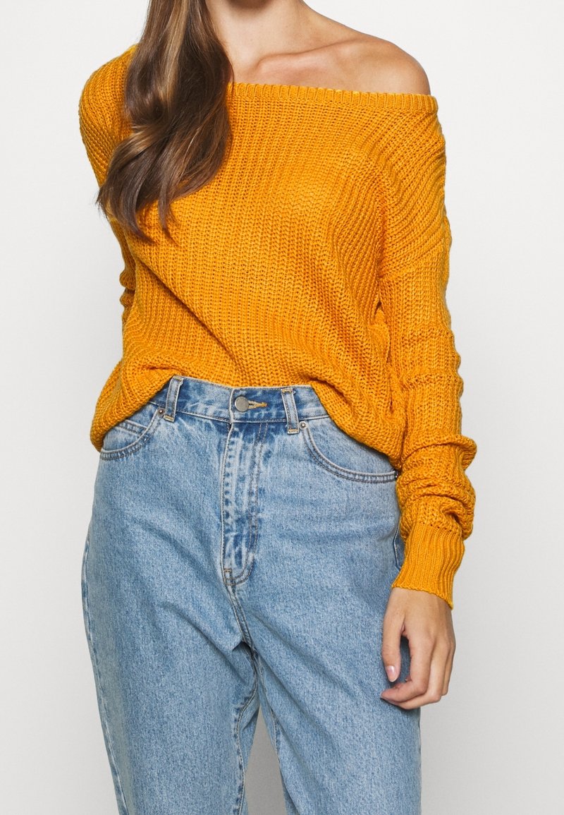 Pull tricoté à épaules dénudées de couleur orange vif, avec une texture côtelée et des manches longues, associé à un jean taille haute bleu clair.
