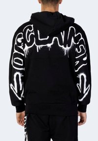 Persona che indossa un felpa nera con testo in stile graffiti bianco e frecce sulla schiena e sulle maniche, abbinata a pantaloni neri coordinati.
