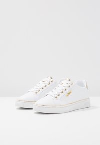 Baskets blanches avec une texture lisse, des accents dorés sur les œillets et le talon, et un logo de marque discret sur le côté. Semelle blanche plate.