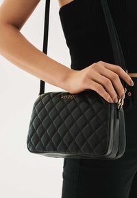 Črna torbica iz prekritega usnja z zlatim napisom "kazar", ramenskim trakom in zadrgo. Oblika je pravokotna s teksturirano površino.