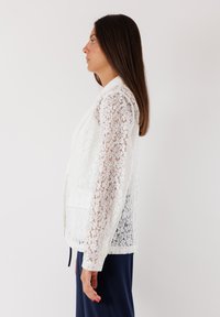 Blazer bianco in pizzo con colletto, presenta maniche in pizzo trasparente, frontale solido e dettagli sulle tasche. Indossato sopra un pantalone blu navy.