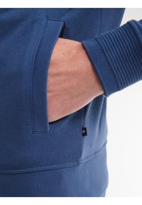 Felpa blu con polsini a coste. Il design presenta una tasca laterale integrata nel corpo e un piccolo etichetta con il marchio vicino all'orlo.