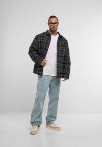 Melnā un pelēkā svītrainā jaka ar pogu aizdari, uzvilkta virs baltas T-krekla. Gaismas zilo džinsu un bežīgu sporta apavu kombinācija noslēdz apģērbu.