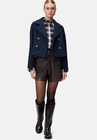 Marineblå dobbeltbrystet cropped jakke, ternet bluse med flæsekrave, brune lædershorts og blanke knæhøje støvler.