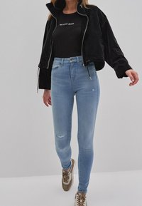 Kvinna som bär ljusblå slitna skinny jeans, svart kort jacka, svart t-shirt med ”Big Star Jeans” och beige sneakers, som går framåt.