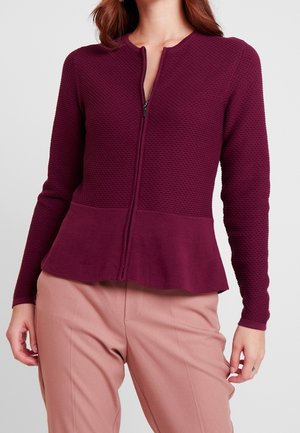 Femme portant une veste zippée texturée bordeaux avec un bas à basque et un pantalon rose clair, debout, les mains détendues le long du corps.