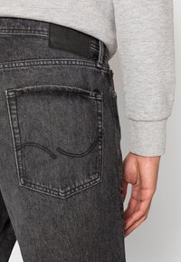 Jeans de mezclilla negros con una superficie texturizada, que presentan un bolsillo trasero con costuras sutiles y un parche rectangular de cuero negro encima de la cinturilla.