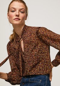 Blusa floreale marrone con scollatura a chiave, maniche ondulate e dettaglio con fiocco. Presenta piccoli motivi di fiori gialli e rossi su un tessuto leggero.