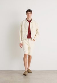 Vaalean kermaisen denim-takin päällä burgundinpunainen t-paita, yhdistettynä kermaisiin shortseihin ja ruskeisiin liukuviin kenkiin. Silkkinen kangas, rento istuvuus.