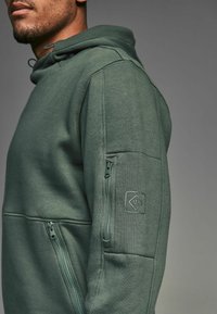 Sudadera verde oscuro con un bolsillo frontal, bolsillo con cremallera en el costado de la manga, tela texturizada y un parche de logo cuadrado en la parte superior del brazo.