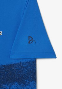 Blå idrottströja med en strukturerad tygkvalitet, med ett gradientmönster från ljusblå till mörkblå, och en logotyp i mörkblått på ärmen.