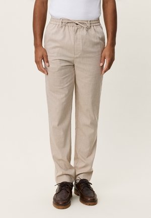 Homme portant un pantalon beige à cordon, une chemise blanche et des chaussures lacées marron foncé, debout devant un fond clair uni.
