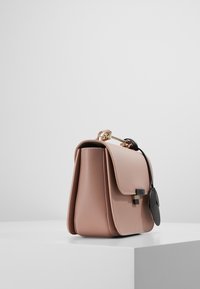 Růžová kožená kabelka se strukturovaným tvarem, zlatými detaily a černým kruhovým štítkem. Hladký povrch a detaily s klopou.