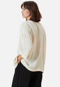 Blusa di colore crema chiaro con vestibilità ampia e maniche a tre quarti, caratterizzata da una texture morbida e tasche laterali. Abbinata a pantaloni neri.