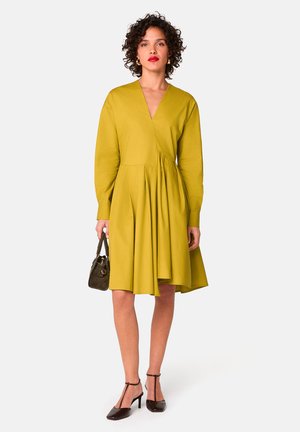 Mosterdkleurige jurk met een V-hals, lange mouwen en een geplooide rok. Gemaakt met donkere hakken en een kleine zwarte handtas.