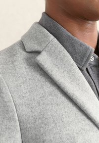 Hallhallane villasegu bleiser, millel on nurgeline krae, tekstureeritud kangas ja tumehall särk all, millel on nööpidega krae.