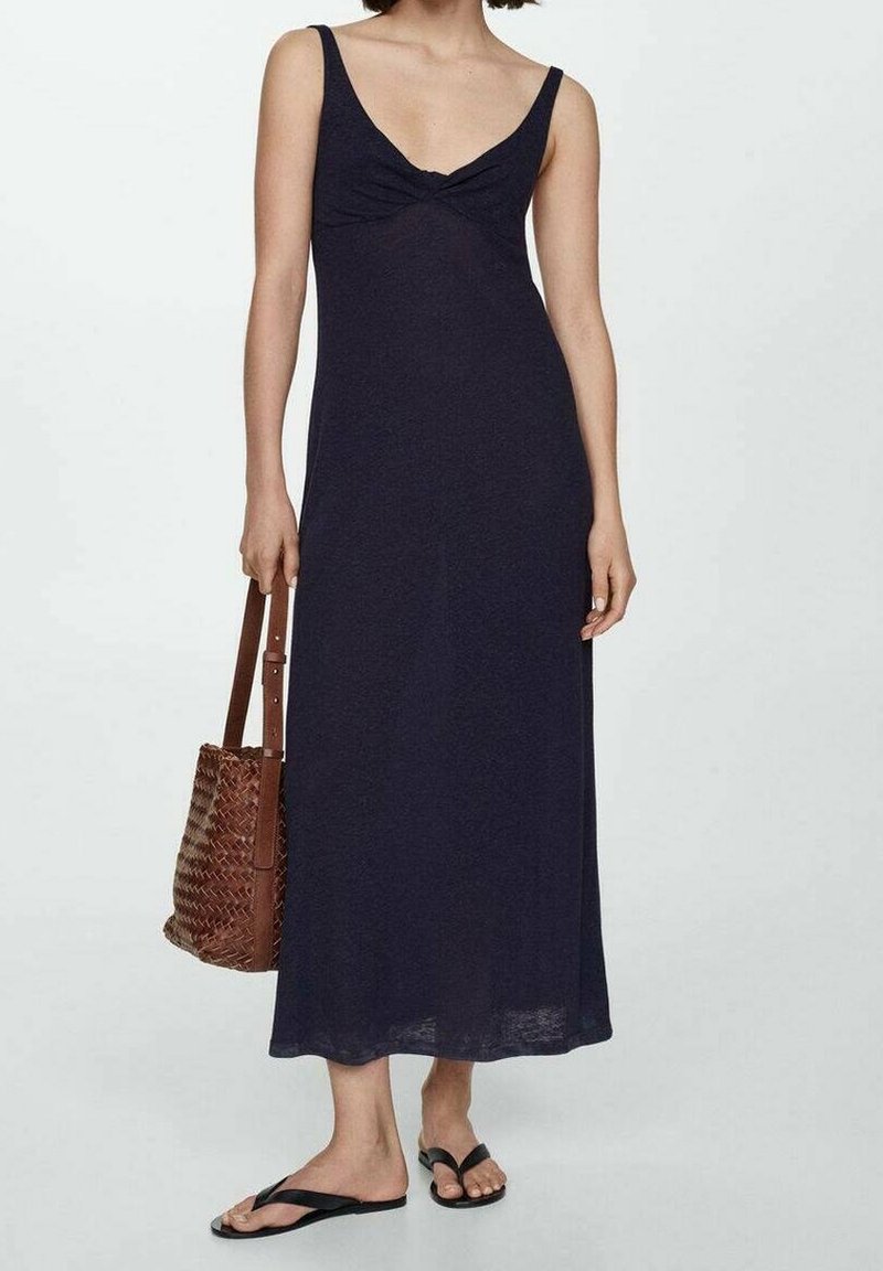 Femme portant une robe midi sans manches bleu marine foncé, tenant un sac à main tressé marron et portant des tongs noires, debout sur un fond blanc.