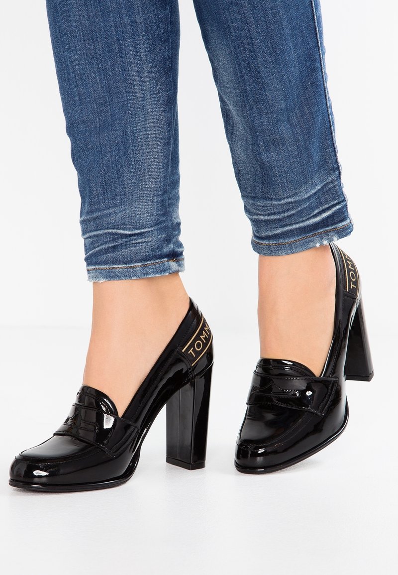 Jambes portant un jean bleu retroussé et des mocassins noirs en cuir verni à talons blocs avec un détail de talon marqué.