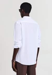 Camicia bianca a maniche lunghe con vestibilità morbida, tessuto texturizzato, polsini con bottoni e orlo curvo. La vista posteriore mostra un sottile dettaglio di yoke.