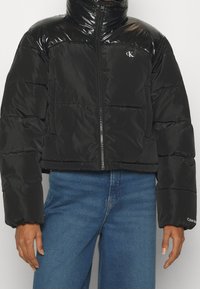 Osoba w czarnej kurtce puchowej z suwakiem, z logo CK na klatce piersiowej, logo Calvin Klein na rękawie i w niebieskich dżinsach, na jednolitym tle.