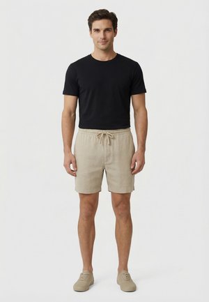 Hombre de pie mirando hacia adelante, vestido con una camiseta negra de manga corta, pantalones cortos beige con cordón y zapatos casuales beige sobre un fondo blanco.