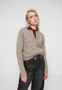 Μπεζ φούτερ ζακέτα με στρογγυλή λαιμόκοψη, κουμπί και μακριά μανίκια, συνδυασμένη με μια σκούρα γκρι ζώνη και μαύρο παντελόνι ψηλής μέσης.