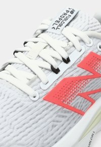 Chaussure de sport gris clair avec une tige texturée, des lacets blancs et un logo corail lumineux sur le côté. Présente un détail de conception perforé.