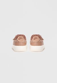 Nike sportcipők világosbarna velúr felsőrésszel, fehér gumitalppal, hatszögletű logóval a saroknál és eltérő varrási részletekkel.