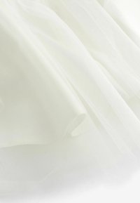Tissu en tulle blanc, léger et transparent, avec une texture douce et fluide. Le matériau tombe élégamment, affichant des plis et des couches délicates.