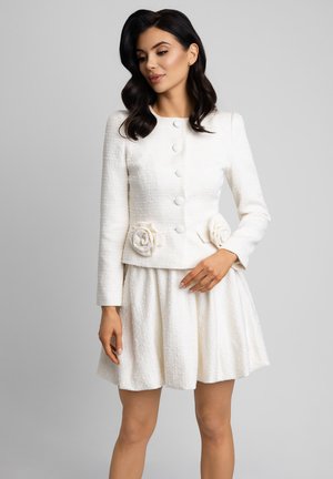 Kobieta o ciemnych, falowanych włosach nosi białą, teksturowaną, zapinaną na guziki kurtkę z kwiatowymi zdobieniami oraz dopasowaną rozkloszowaną spódnicę na jednolitym szarym tle.