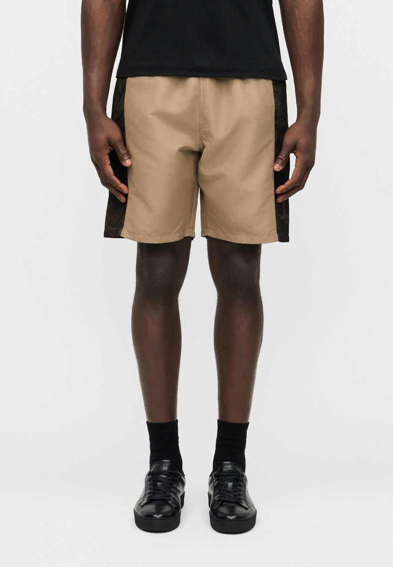 Homme portant un short beige avec des panneaux latéraux en mesh noir, des chaussettes noires, des chaussures noires et une chemise noire, debout devant un fond blanc.