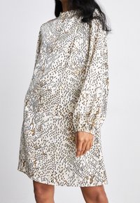 Robe crème à manches longues avec un motif abstrait texturé en noir et doré, dotée d'une encolure haute et de poignets de manches froncés.