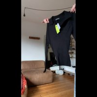 Svart langermet skjorte med kragehals, ribbet tekstur og et grønt prislapp festet. Bakgrunnen har en brun sofa og hyller.