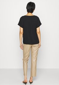 Personne avec des cheveux courts et foncés, portant un haut noir ample à manches courtes, un pantalon beige slim et des sandales à talons noires, se tenant devant un fond blanc.