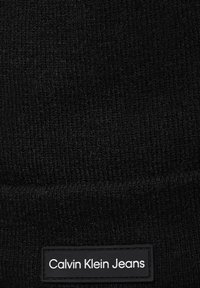 Fekete kötött sapka textúrált felülettel. Jellemzője egy téglalap alakú szövet címke, rajta "Calvin Klein Jeans" felirattal fehér betűkkel, fekete háttéren.