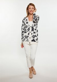 Cardigan a maglia di colore crema con motivi floreali neri, dotato di scollatura a V, chiusura con bottoni e polsini a coste; abbinato a pantaloni bianchi e mocassini beige.