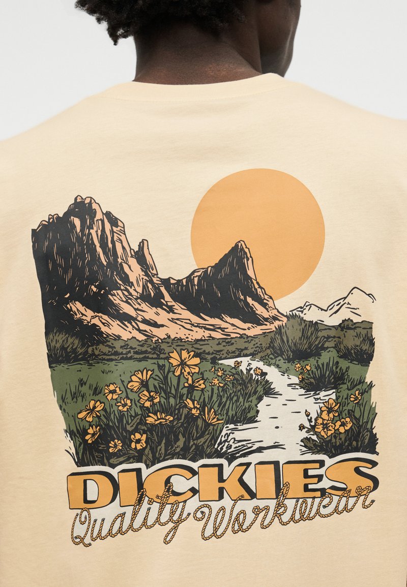 Person trägt ein beiges Hemd mit einer Grafik von Bergen, Wildblumen, Sonne und dem Text "DICKIES Quality Workwear" auf der Rückseite.