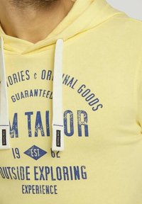 Felpa con cappuccio gialla con cordini bianchi, con testo blu che include "Tom Tailor", "Outside Exploring" e un design grafico vintage.