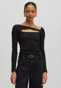 Schwarzes Langarmoberteil mit geripptem Textil, das über ein diagonales Ausschnittdetail und eine schmale Passform verfügt. Kombiniert mit dunkler Hose und einem Gürtel.
