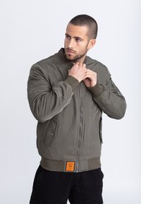 Hombre con el pelo corto que lleva una chaqueta bomber verde oliva, subiendo la cremallera del cuello, mirando de lado sobre un fondo gris liso.