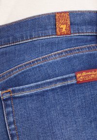 Primer plano de pantalones vaqueros de mezclilla azul que muestran el bolsillo trasero, costuras naranjas y etiquetas de la marca rojas con letras amarillas.