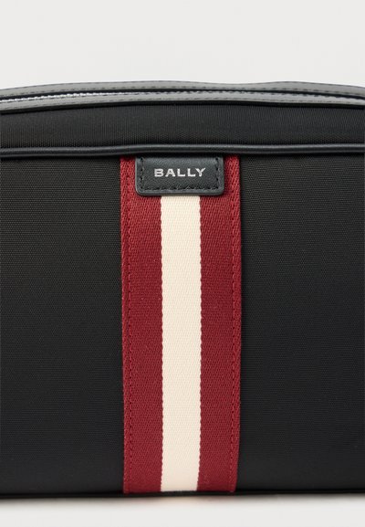 Μαύρη υφασμάτινη τσάντα με μια κεντρική κάθετη κόκκινη και λευκή ρίγα και ένα μικρό μαύρο δερμάτινο λογότυπο με την επιγραφή "BALLY" στην κορυφή στο κέντρο.