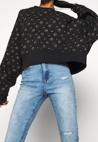 Sweat-shirt noir avec motif de logo circulaire doré, coupe oversize, poignets côtelés. Associé à un jean en denim bleu clair avec des détails usés.