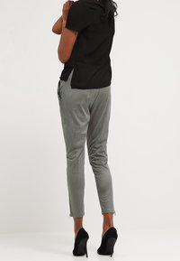 Zwarte t-shirt met een doorschijnende achterkant, grijze slim-fit broek met een gladde textuur, en zwarte hoge hakken. Achteraanzicht toont de details van de outfit.