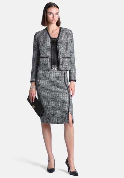 Femme portant un tailleur jupe gris texturé avec une bordure noire, un haut noir, tenant une pochette noire et portant des talons hauts noirs.