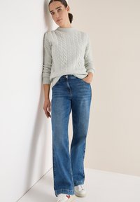 Lichtgrijze kabelgebreide trui met geribbelde halslijn, gecombineerd met donkerblauwe flared jeans en witte sneakers. Gladde textuur en een ontspannen pasvorm.