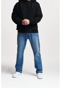 Sudadera negra con cremallera y bolsillos frontales, combinada con jeans rectos azules y zapatillas blancas. Tejido suave y diseño holgado.