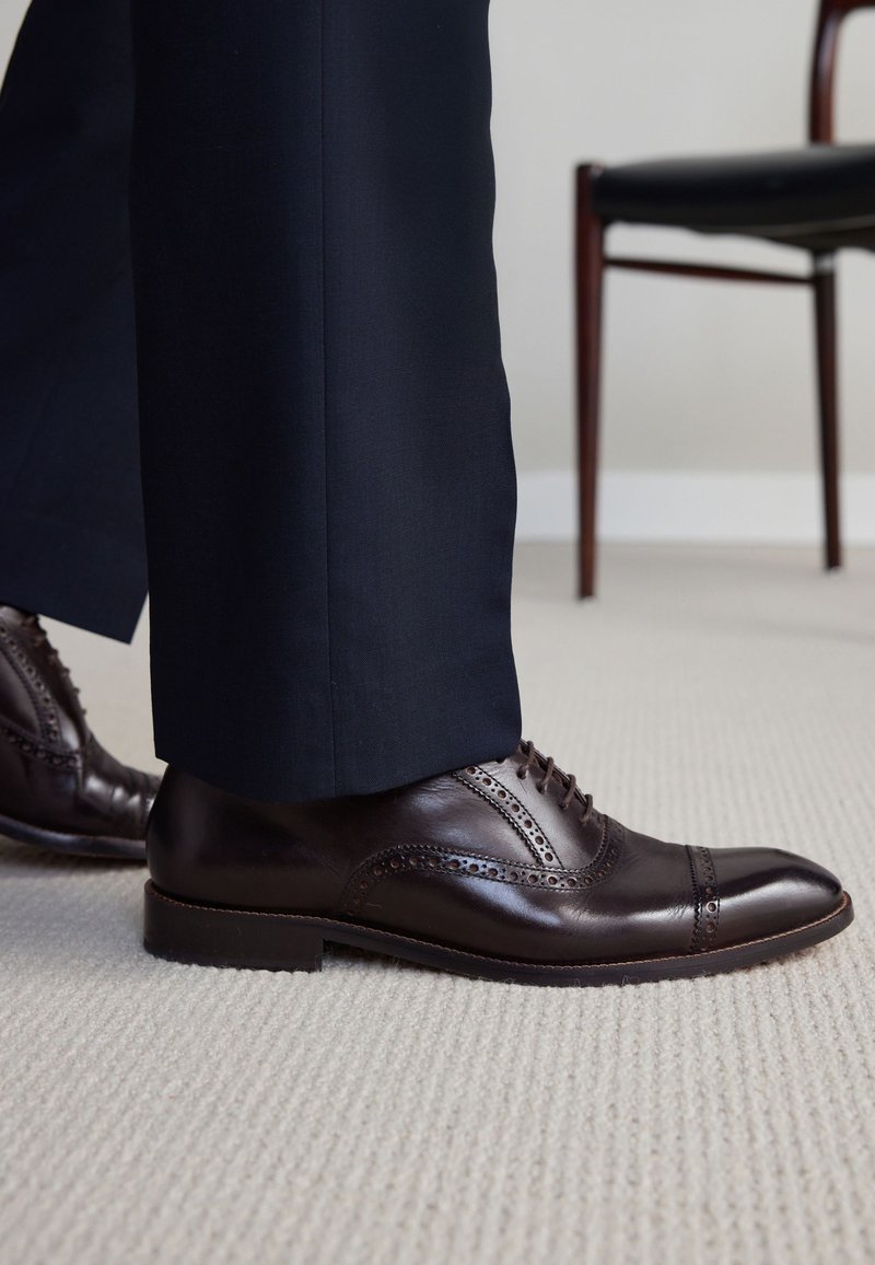 Chaussures habillées en cuir marron avec des détails en brogue portées avec un pantalon bleu marine foncé sur un tapis clair, chaise en bois partiellement visible en arrière-plan.
